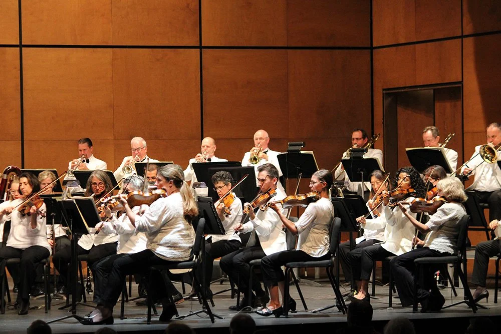 An orchestra performing on stage with musicians playing violins, trumpets, and other instruments, against a wooden paneled background.