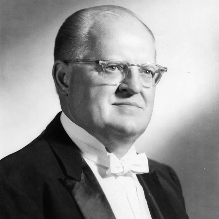 Black and white portrait of a man wearing glasses, a tuxedo, and a bow tie.