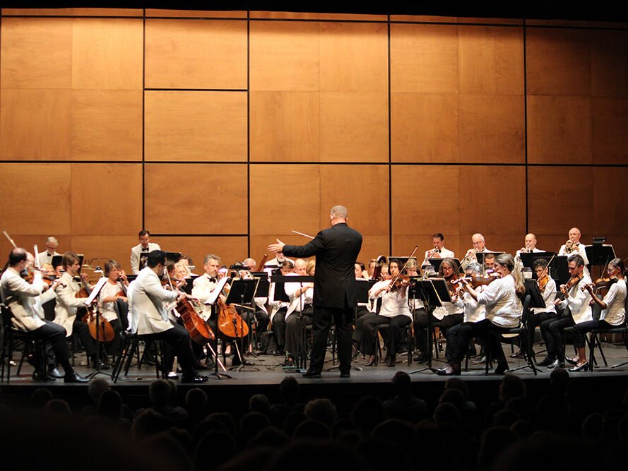 An orchestra performing on stage with a conductor leading them, in front of a wooden-paneled wall.