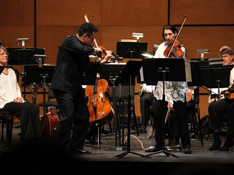 Orchestra performing on stage with musicians playing violins, a cello, and other string instruments.