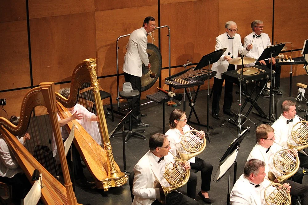 Orchestra performing on stage with musicians playing harp, French horns, percussion instruments, and brass instruments, all dressed in formal white shirts and black bow ties.
