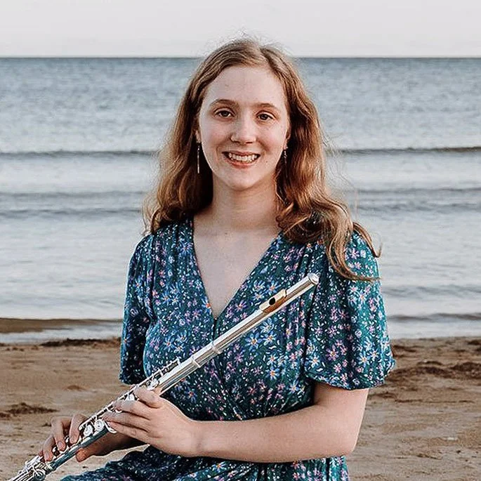 A woman with light skin, long wavy hair, and earrings standing on a beach holding a flute, with the ocean in the background.