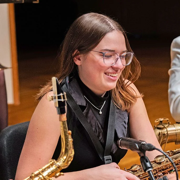 Young woman wearing glasses, smiling, sitting at a table with saxophones, microphone, and music stand, possibly in a music rehearsal or performance setting.