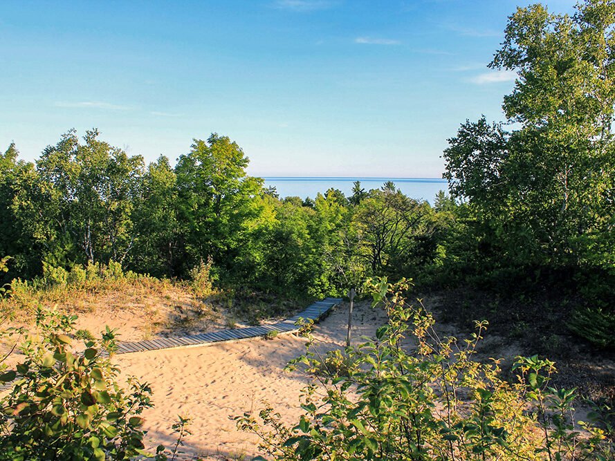 A sandy pathway with a wooden walkway snaking through green trees leading to a distant body of water under a blue sky.