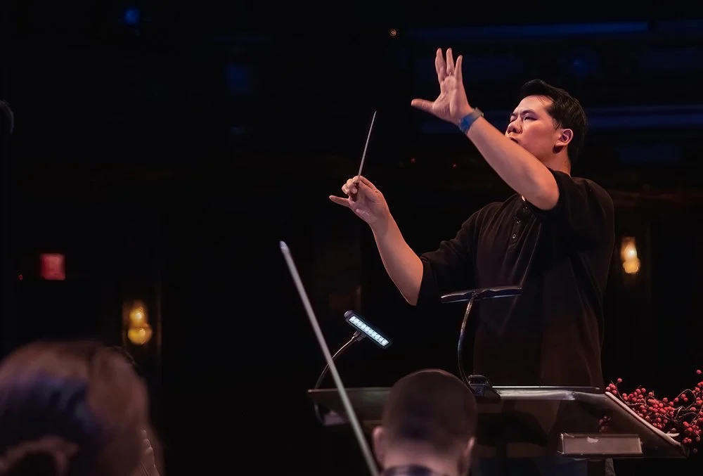 A woman standing at a conductor's podium, conducting an orchestra, with her right hand raised and holding a baton, and her left hand in motion, set against a dark background with dim lighting.