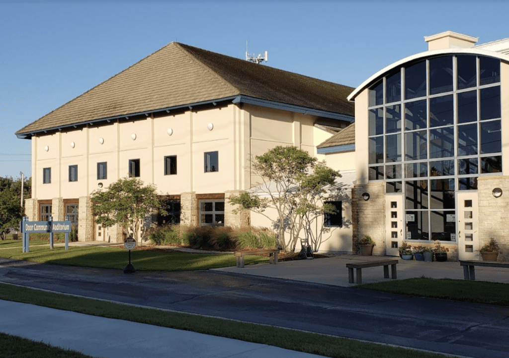 A large building with a sign that reads "Door Community Auditorium" in front of it, featuring beige walls, a gabled roof, and a glass entrance with arched windows, surrounded by trees and a lawn.