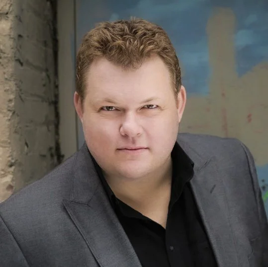 A man with curly blond hair wearing a dark blazer and black shirt, standing indoors against a colorful abstract background.