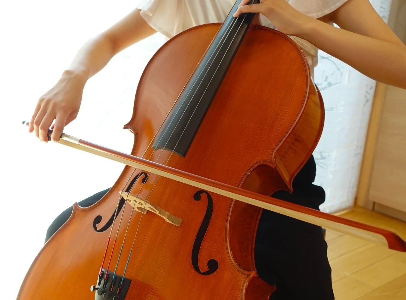 Person playing a wooden cello, holding a bow, with a background of light-colored brick wall and wooden floor.