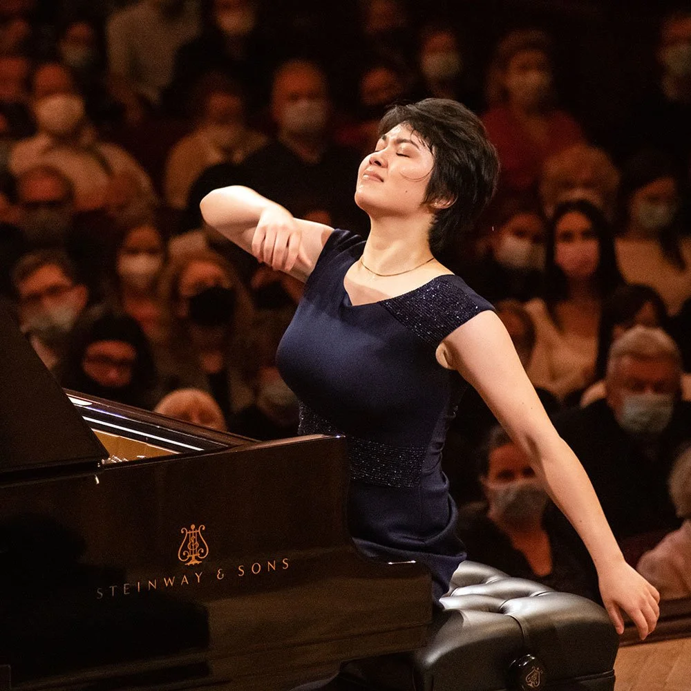 A woman passionately plays a grand piano during a performance with an audience wearing masks in the background.