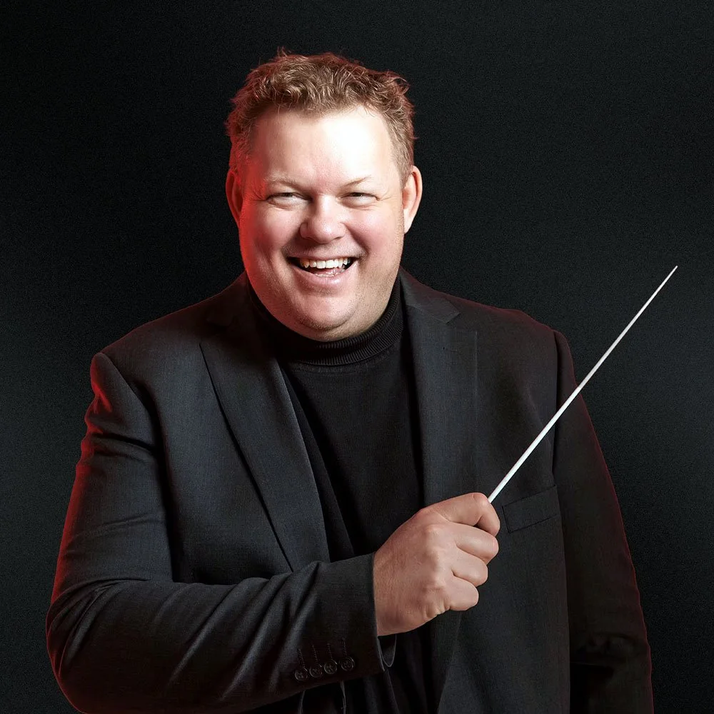 A smiling man with curly red hair, wearing a black blazer and black turtleneck, holding a conductor's baton against a dark background.