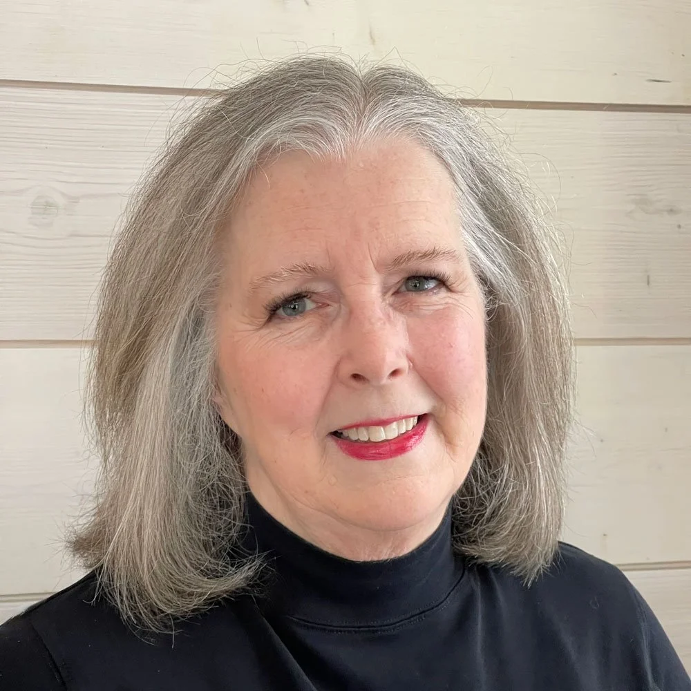 A portrait of an older woman with shoulder-length gray hair, light skin, blue eyes, wearing red lipstick and a black turtleneck, standing against a light-colored wooden wall.