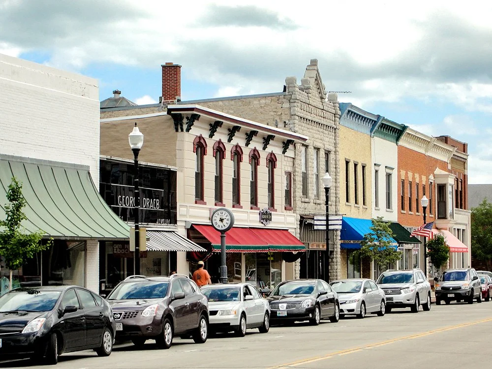 A small town with historic brick and stone storefront buildings, parked cars along the street, and a lamppost with a clock.