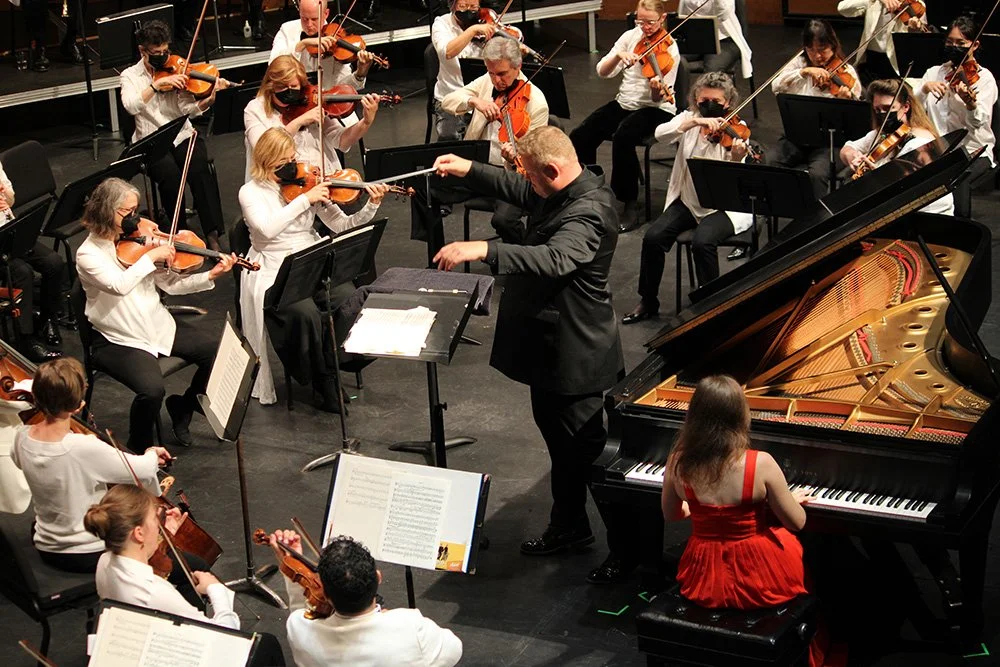 An orchestral concert with musicians playing string instruments and a pianist performing on stage, led by a conductor.