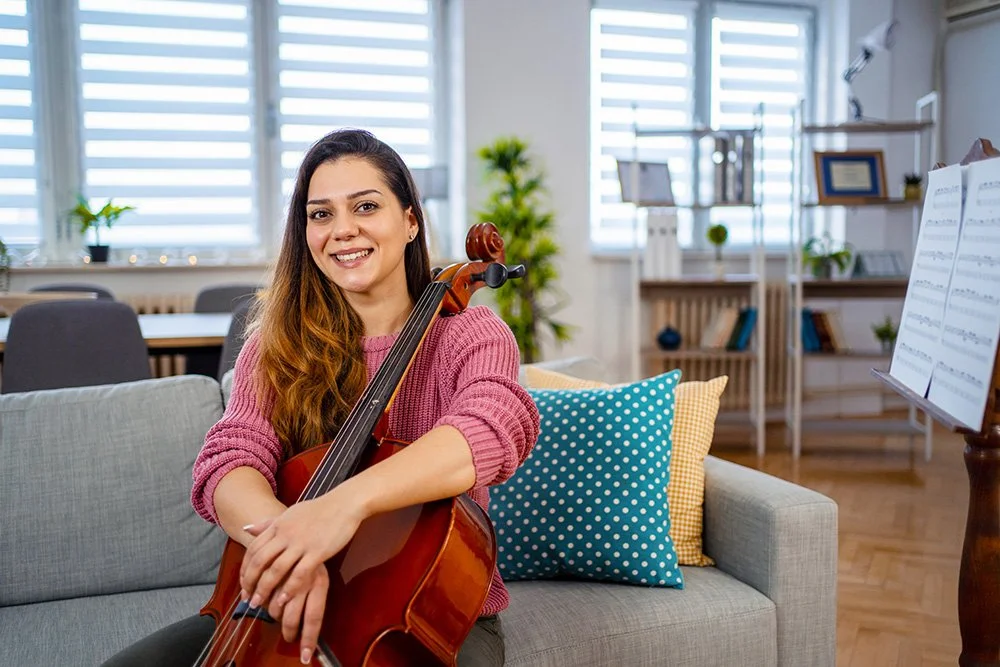 A woman sitting on a couch in a living room, holding a cello and smiling at the camera, with musical sheets on a stand nearby.
