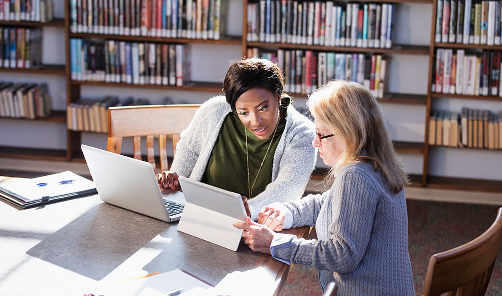 Two women sit at a table in a library, looking at a tablet together with a laptop and books nearby.
