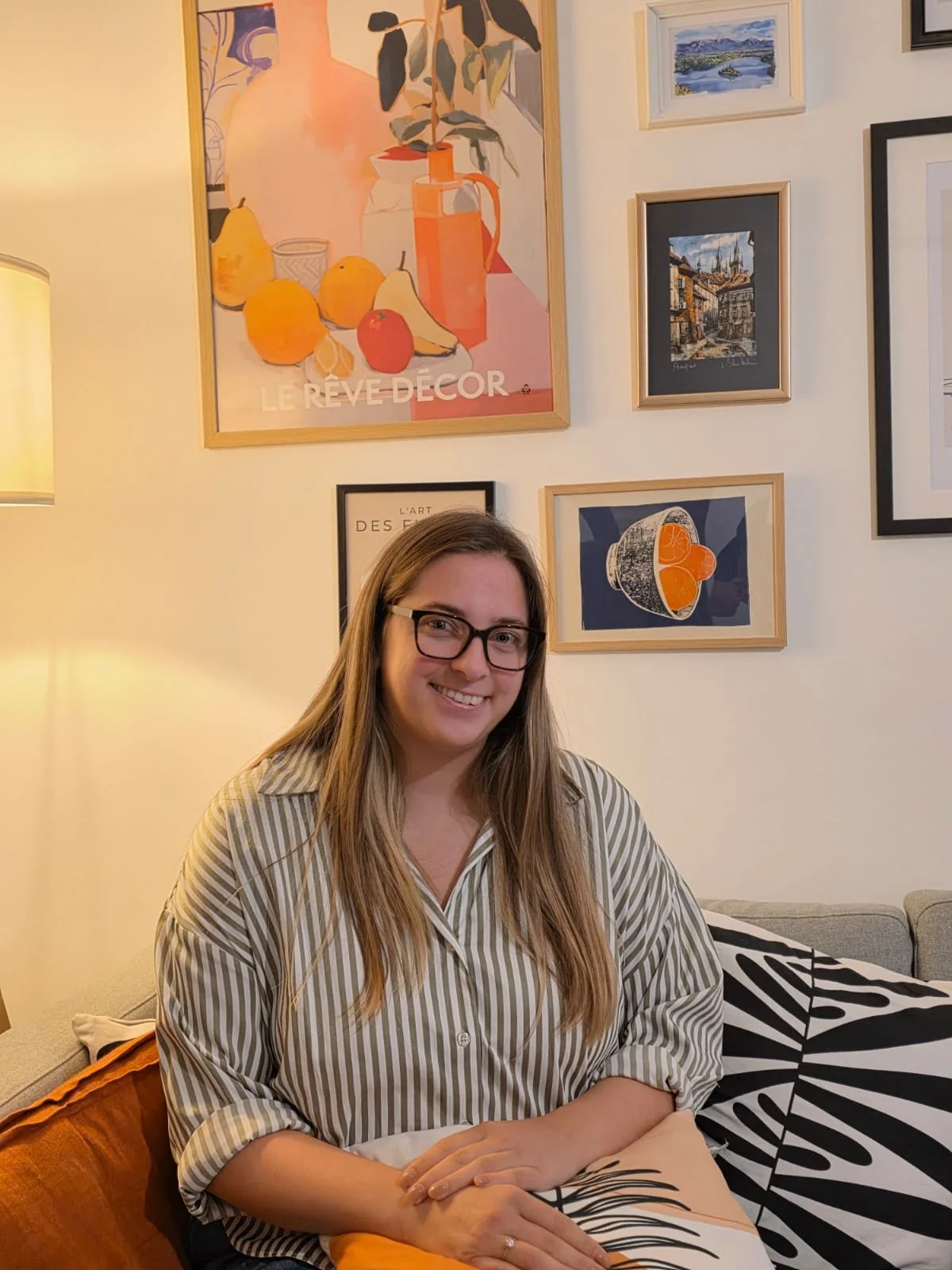 A woman with long blonde hair, wearing glasses and a striped shirt, smiling and sitting on a sofa in a living room with framed artwork on the wall behind her.