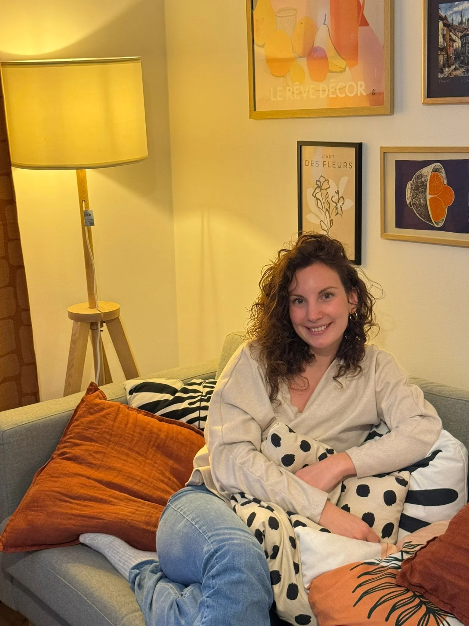 A young woman with curly brown hair, smiling, sitting on a light-colored sofa with pillows. She is wearing a beige shirt and light jeans, holding a polka dot pillow. There is a floor lamp and framed artwork on the wall behind her.