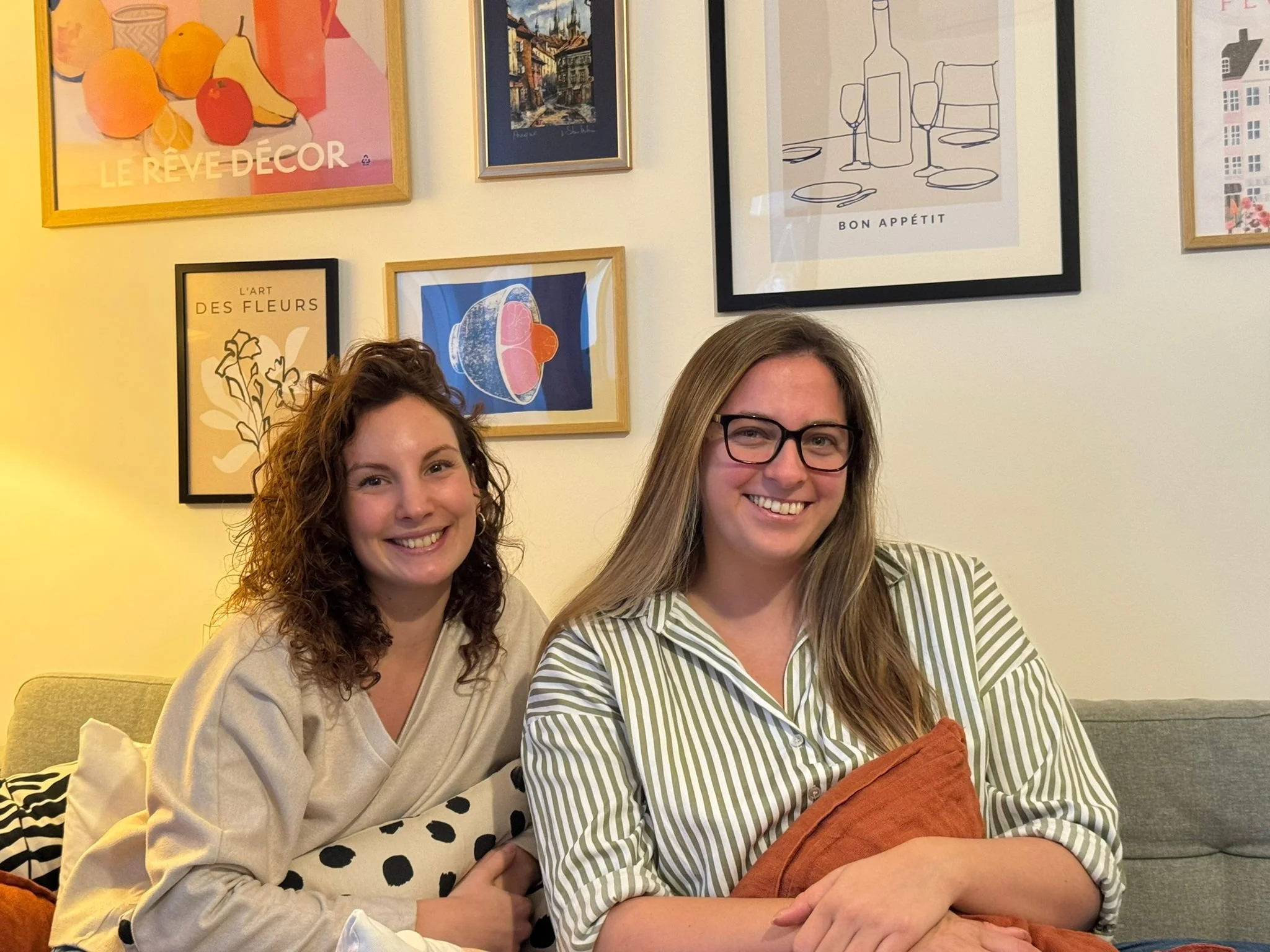 Two smiling women sitting on a couch in a room with framed art on the wall behind them.