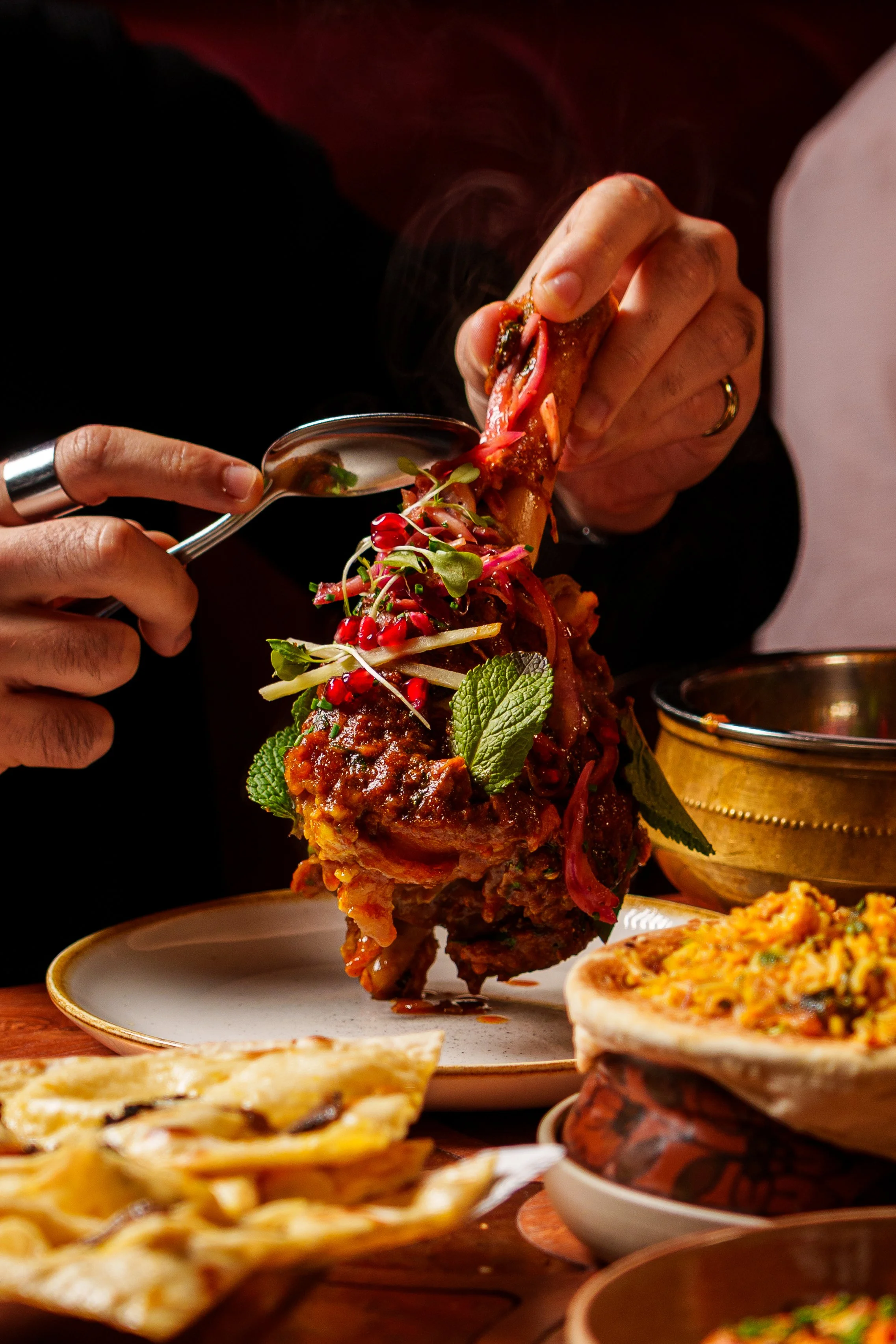 A person is holding a large lamb shank and garnishing it with herbs and microgreens, with various dishes and flatbread on the table.