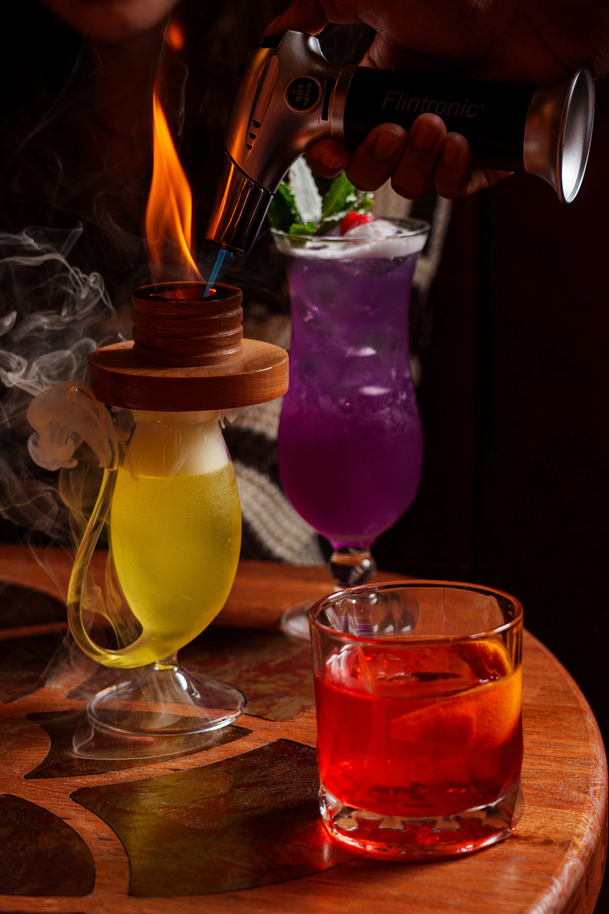 A person is holding a torch flame lighter over a cocktail, with smoke rising around. Two colorful cocktails, one purple and one red, are on a wooden table in the background.