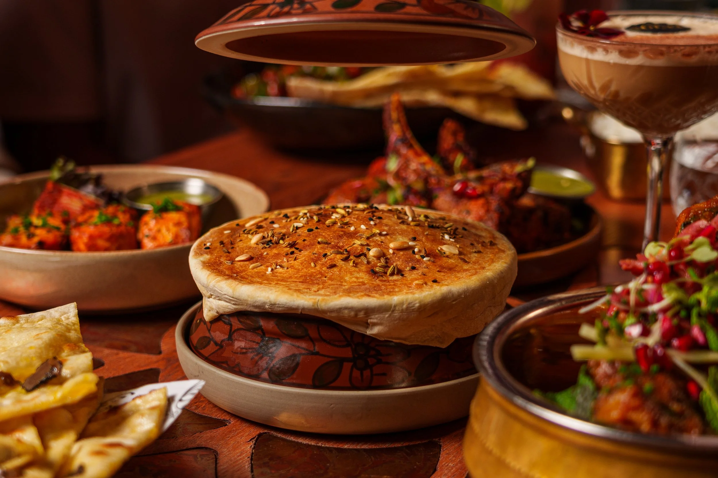 A variety of Indian dishes on a wooden table, including biryani topped with seeds in the center, and other dishes with spices and garnishes.
