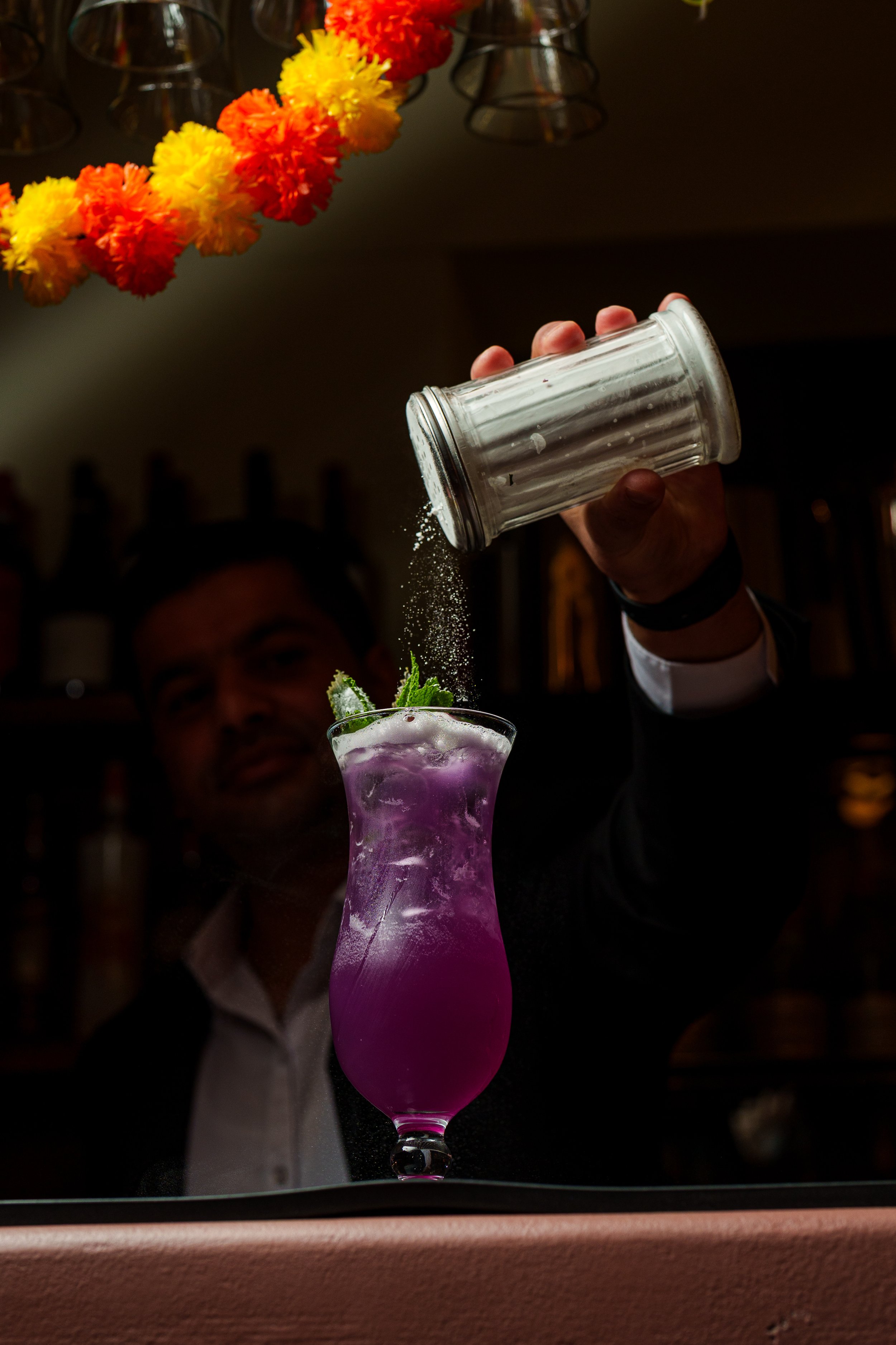 Bartender sprinkling powdered sugar into a purple cocktail garnished with mint and lime.