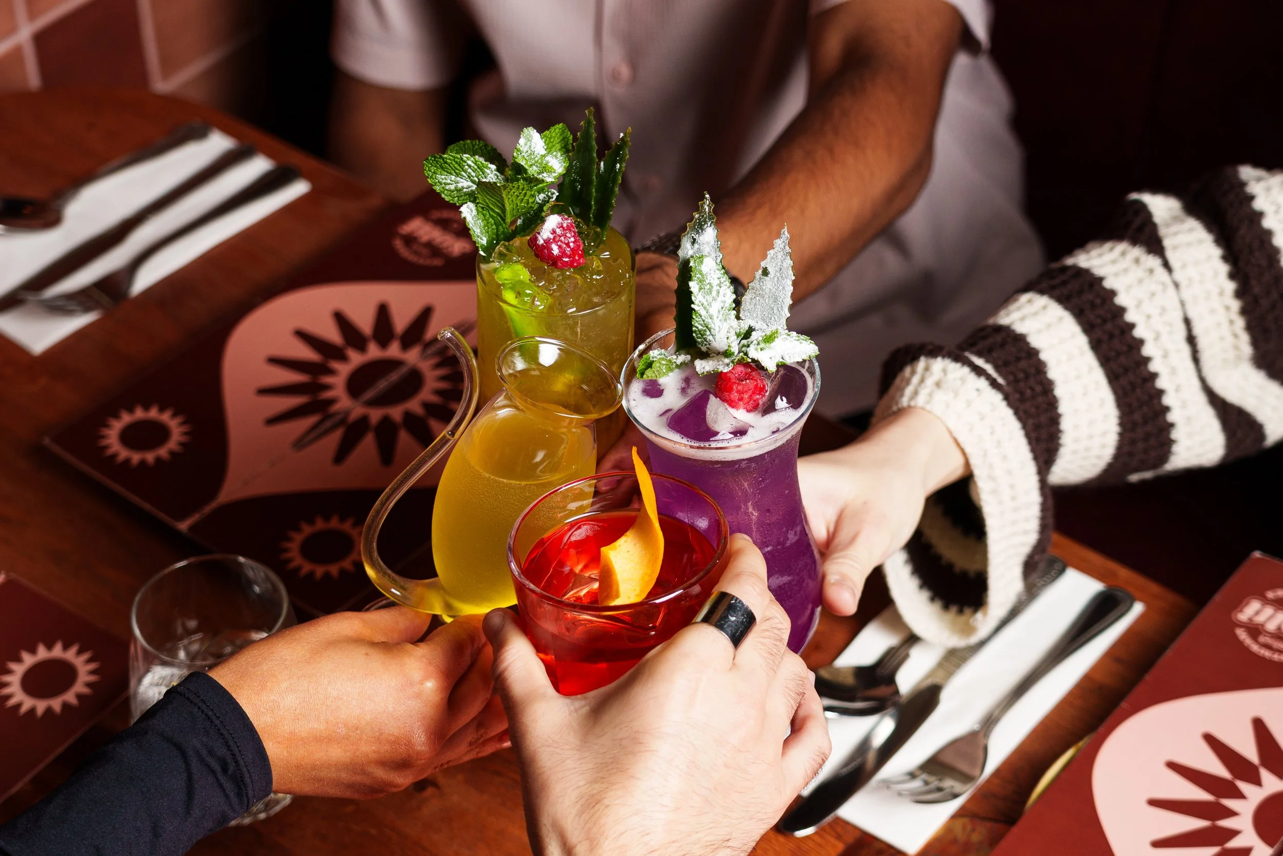Friends clinking colorful cocktails at a restaurant table.