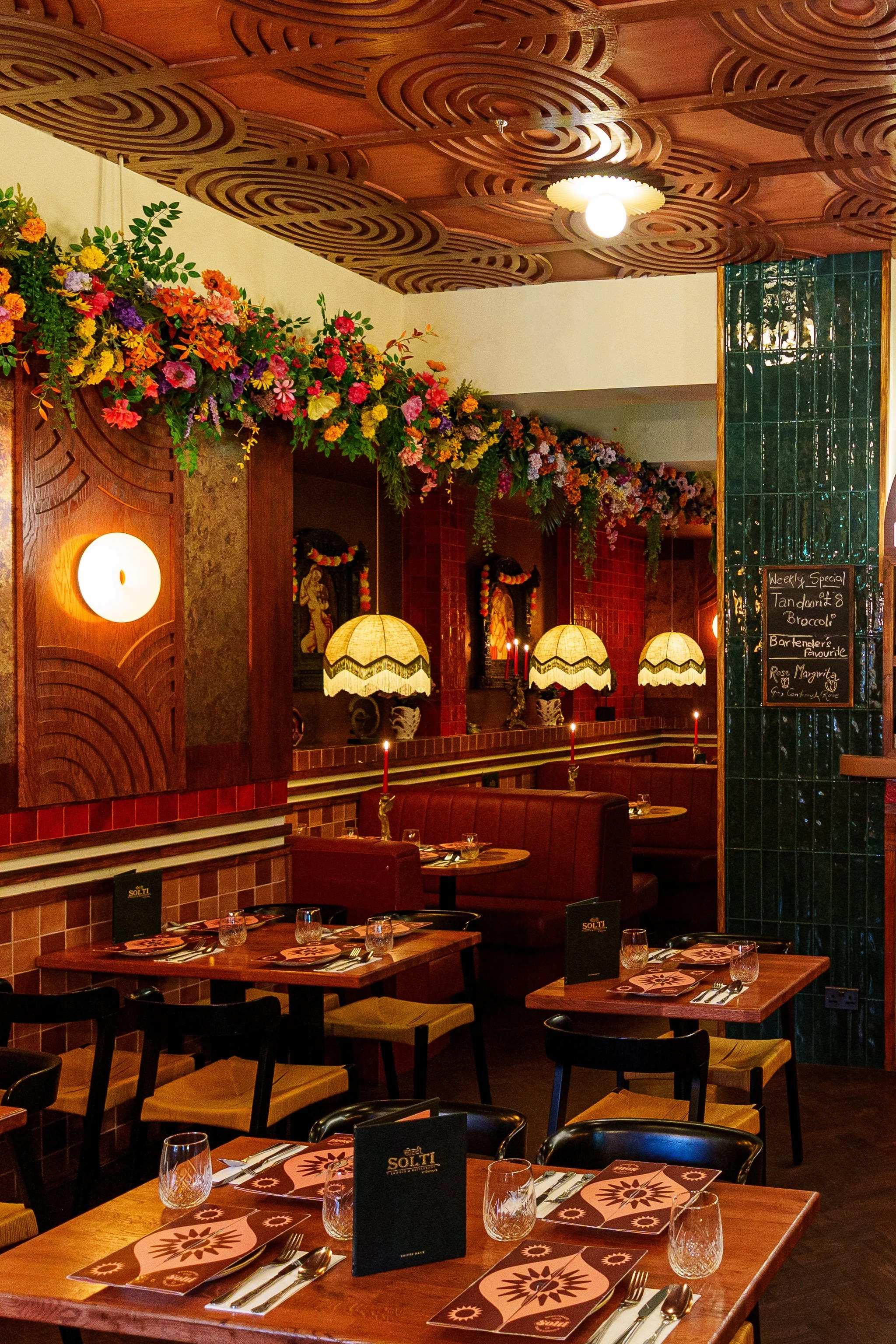 Interior of Solti restaurant with wooden tables set with glasses, napkins, and cutlery, decorated with floral arrangements along the ceiling, with lamps hanging down, and a chalkboard menu on a green tiled wall.