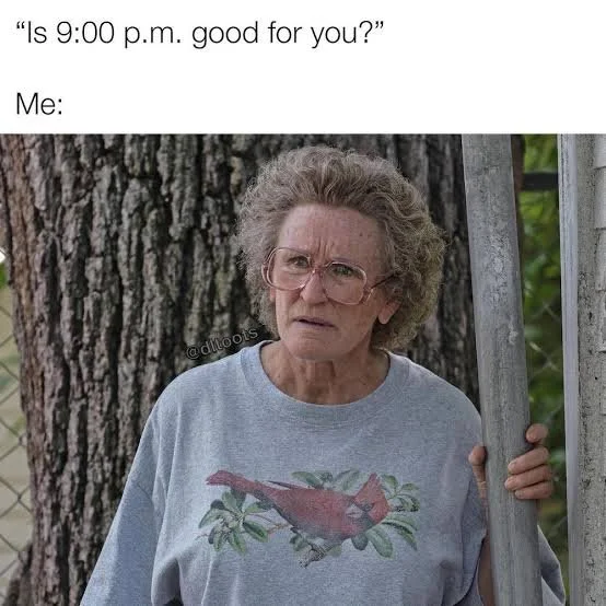 A confused elderly woman with glasses and curly hair stands outside, holding onto a wooden post. She wears a gray T-shirt with a bird graphic and appears puzzled. The background shows a large tree trunk and some green foliage.