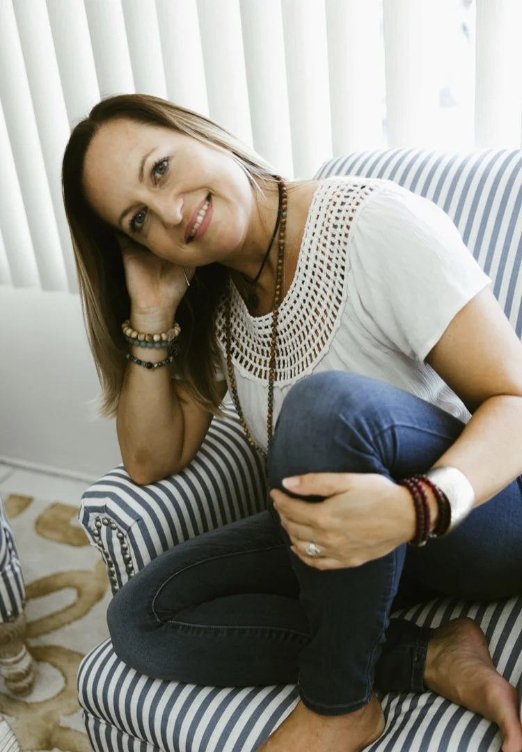 A woman sitting on a striped couch, smiling, with her head resting on her hand. She is wearing a white top with crochet details, jeans, and multiple bracelets and necklaces.