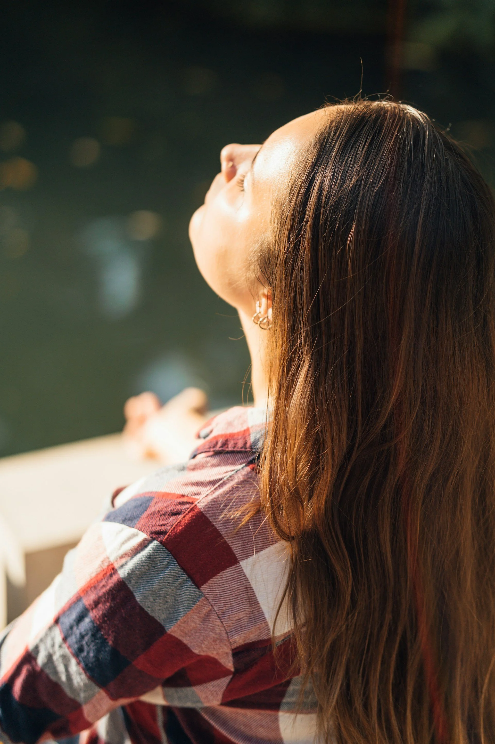 A woman with long brown hair and earrings, wearing a red and white plaid shirt, sitting outdoors with her eyes closed and face tilted upward, in sunlight.