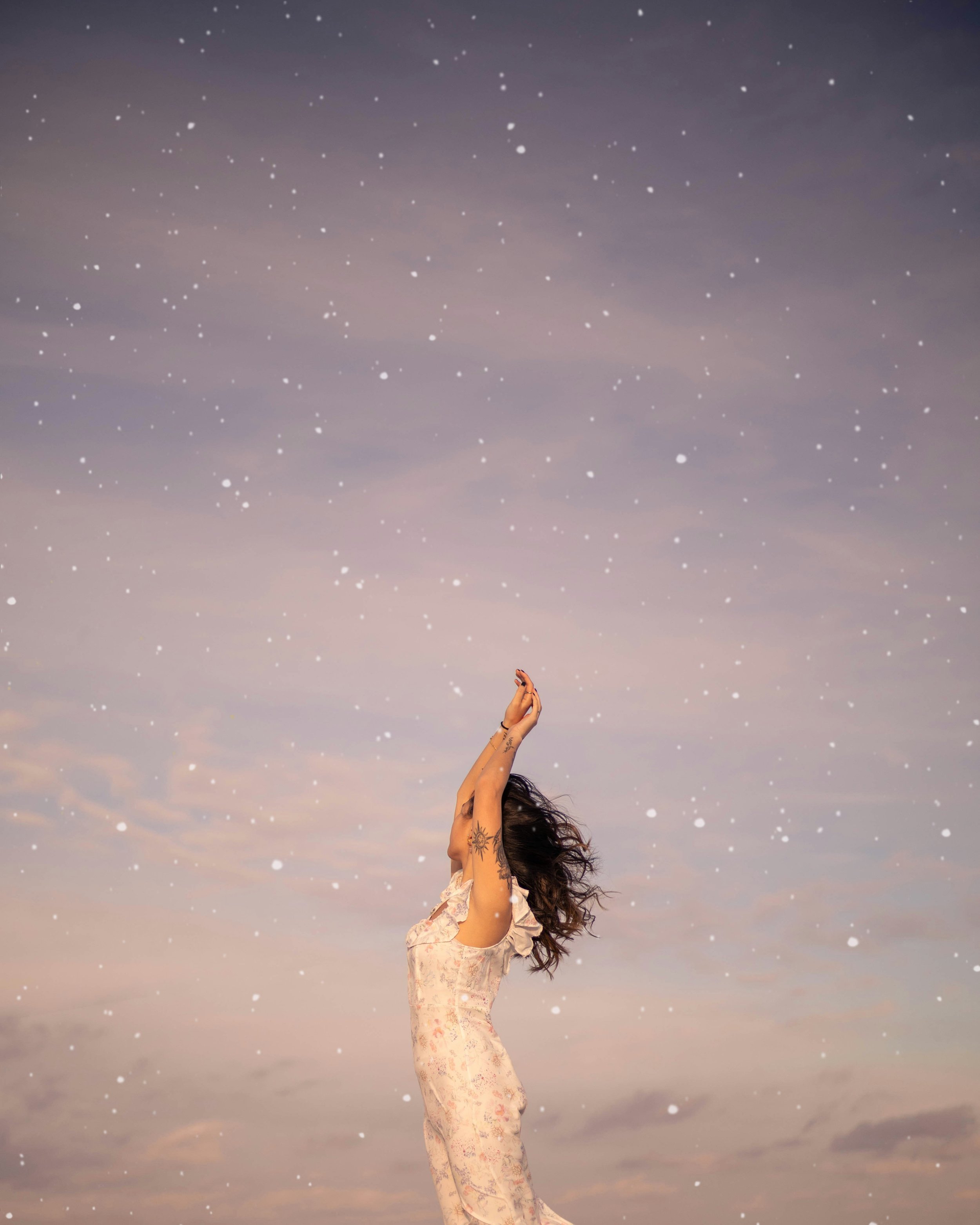 A woman with tattoos on her arms and dark hair flowing, standing outdoors with her arms raised above her head, against a sky with clouds and falling snow or confetti.