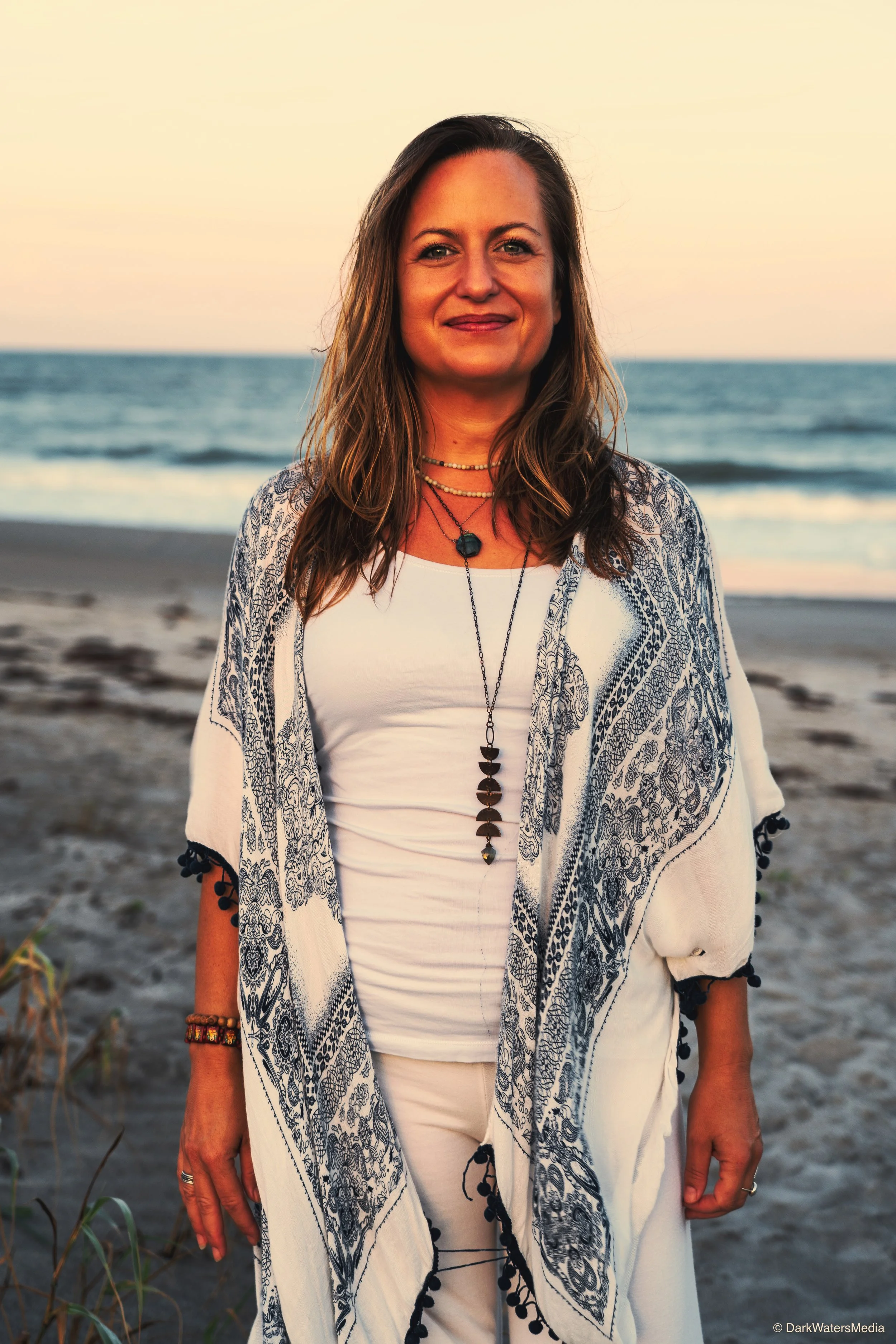 Woman standing on the beach during sunset, facing the camera with a slight smile, wearing a white top and a patterned shawl, with ocean waves in the background.