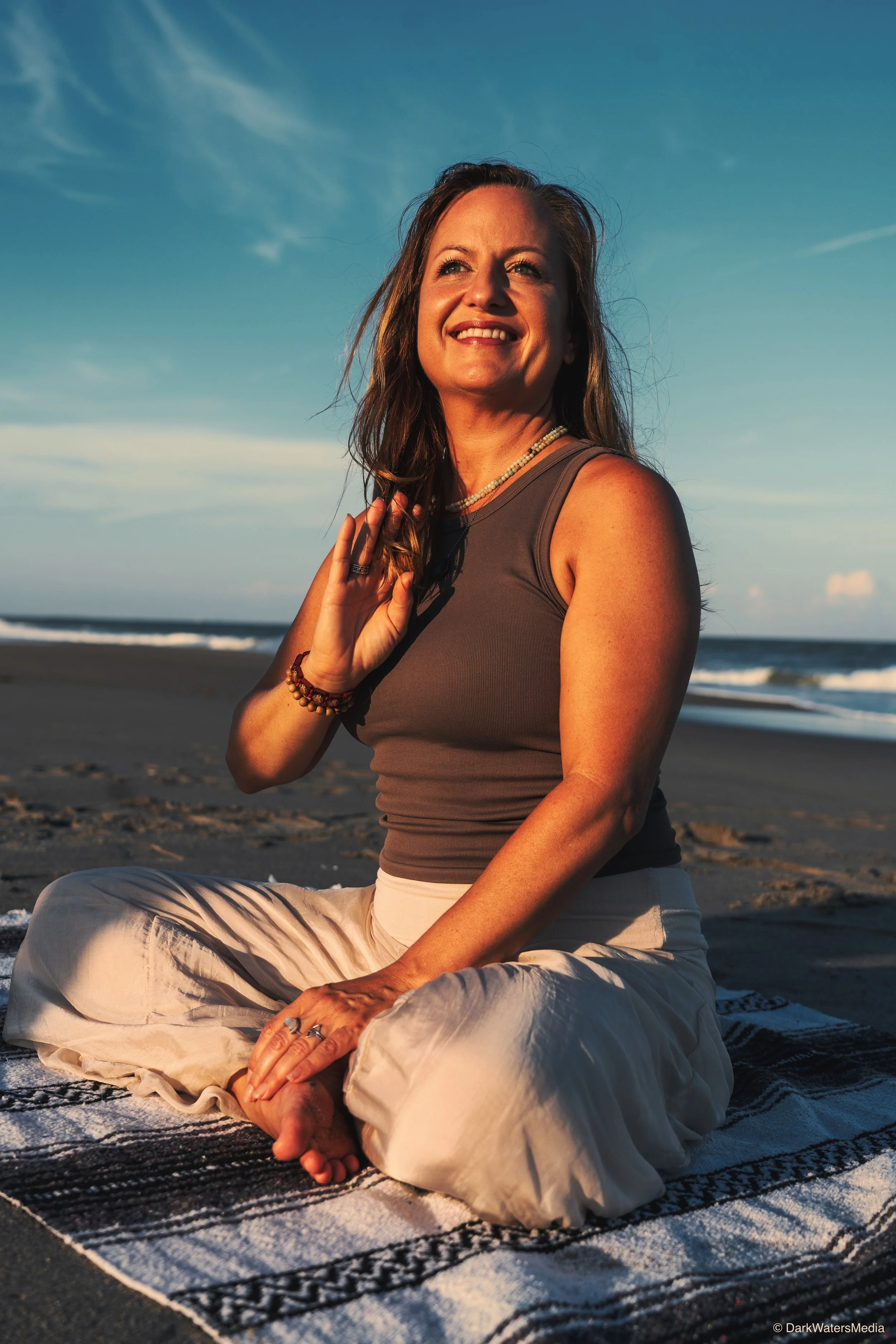 A woman practicing yoga on a beach during sunset, sitting cross-legged on a patterned mat, smiling and holding a hand in a mudra gesture, with waves and a blue sky in the background.