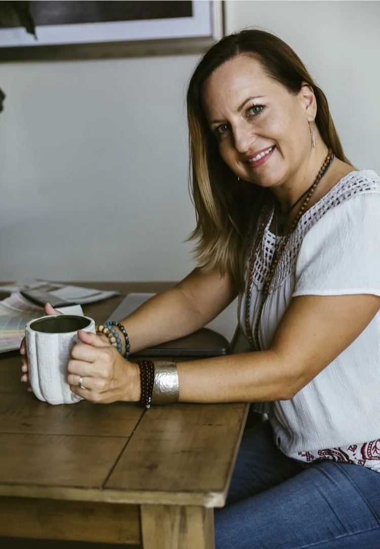 A woman sitting at a wooden table, holding a mug, smiling at the camera, with a laptop and papers in the background.