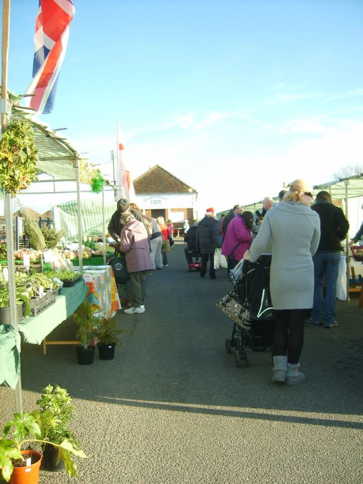People shopping at an outdoor farmers market with stalls under tents and a cafe in the background, sunny day with clear blue sky.