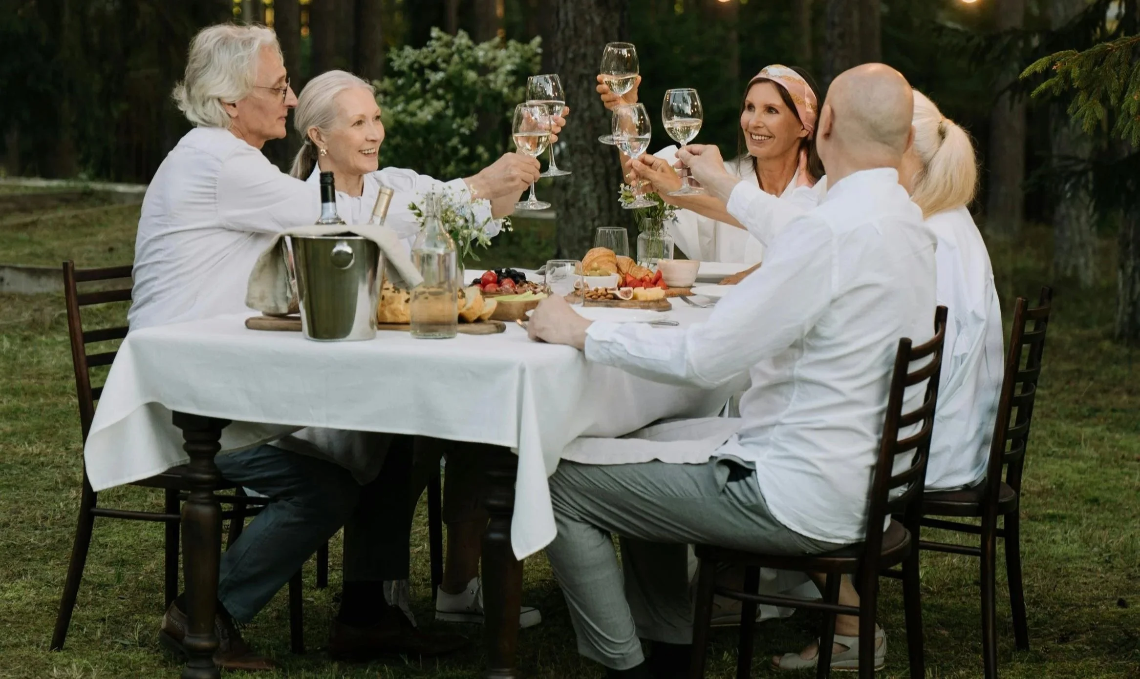A group of six elderly people celebrating outdoors at a dinner table with glasses of wine, surrounded by trees, with food and flowers on the table.