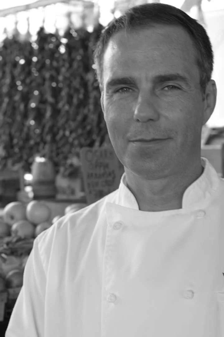 Black and white photo of a male chef in a white coat outdoors near fruits and plants.