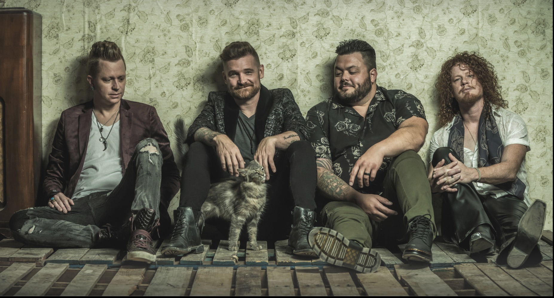 Four men sitting against a floral patterned wallpaper, with a cat in front of the second and third men. They are dressed in casual, slightly edgy clothing and are seated on a platform made of wooden planks.
