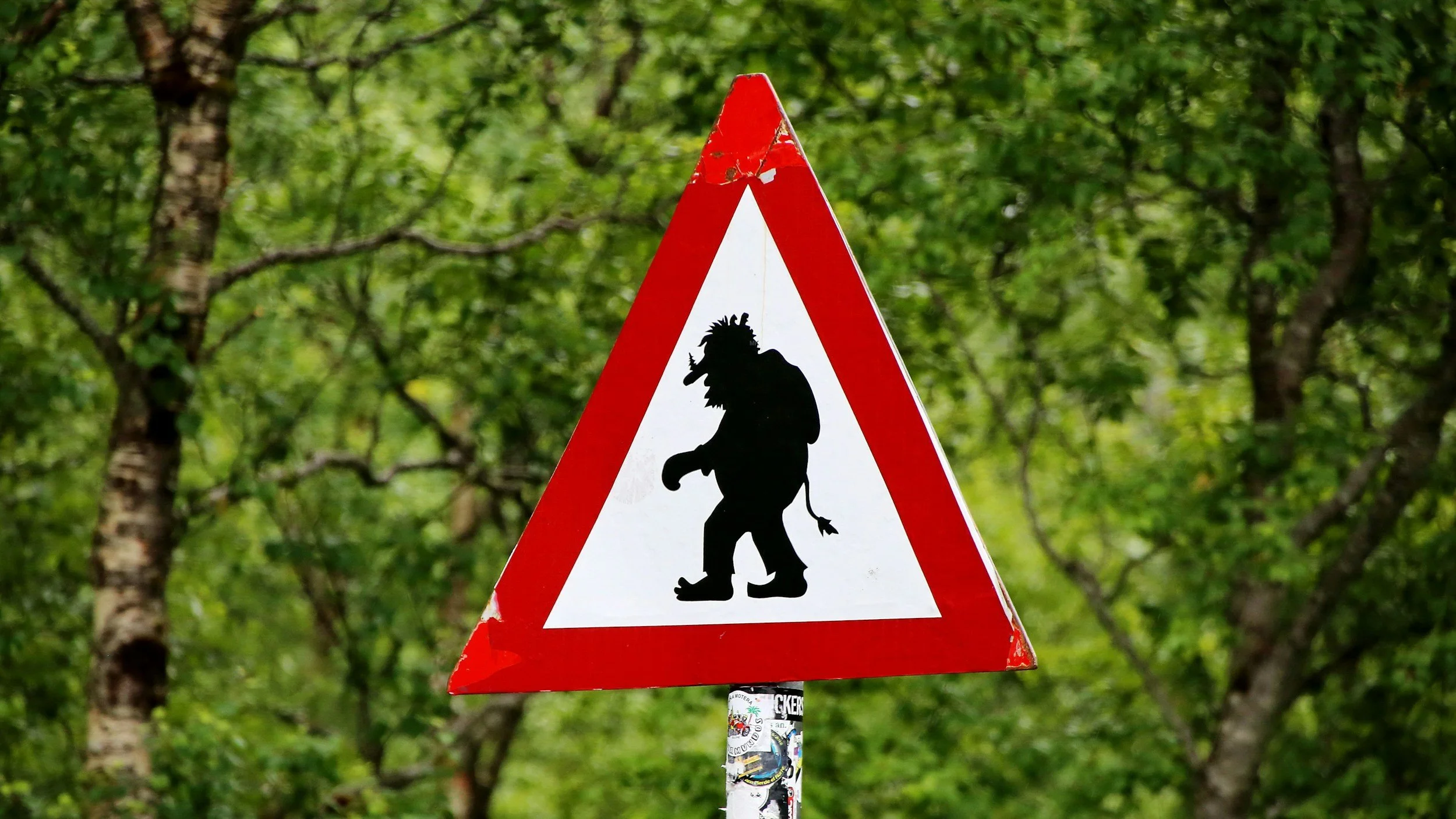 Triangular warning sign with a silhouette of a troll in the center, set against a background of green trees and foliage.