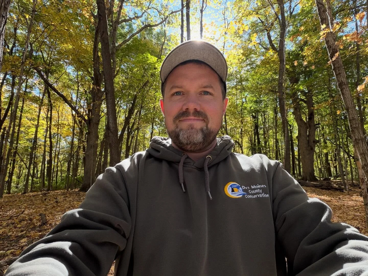 A man taking a selfie in a forest during daytime, wearing a gray hoodie with a logo that reads 'Das Moines County Conservation' and a gray cap. Sunlight filters through the green and yellow autumn leaves.