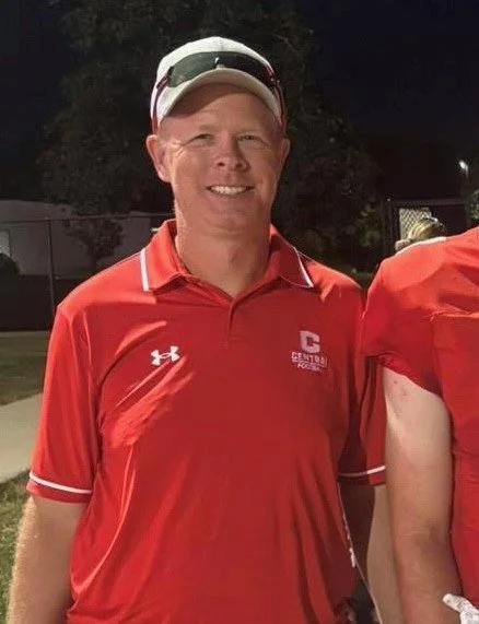 A man in a red shirt and white hat standing at a college football field at night.