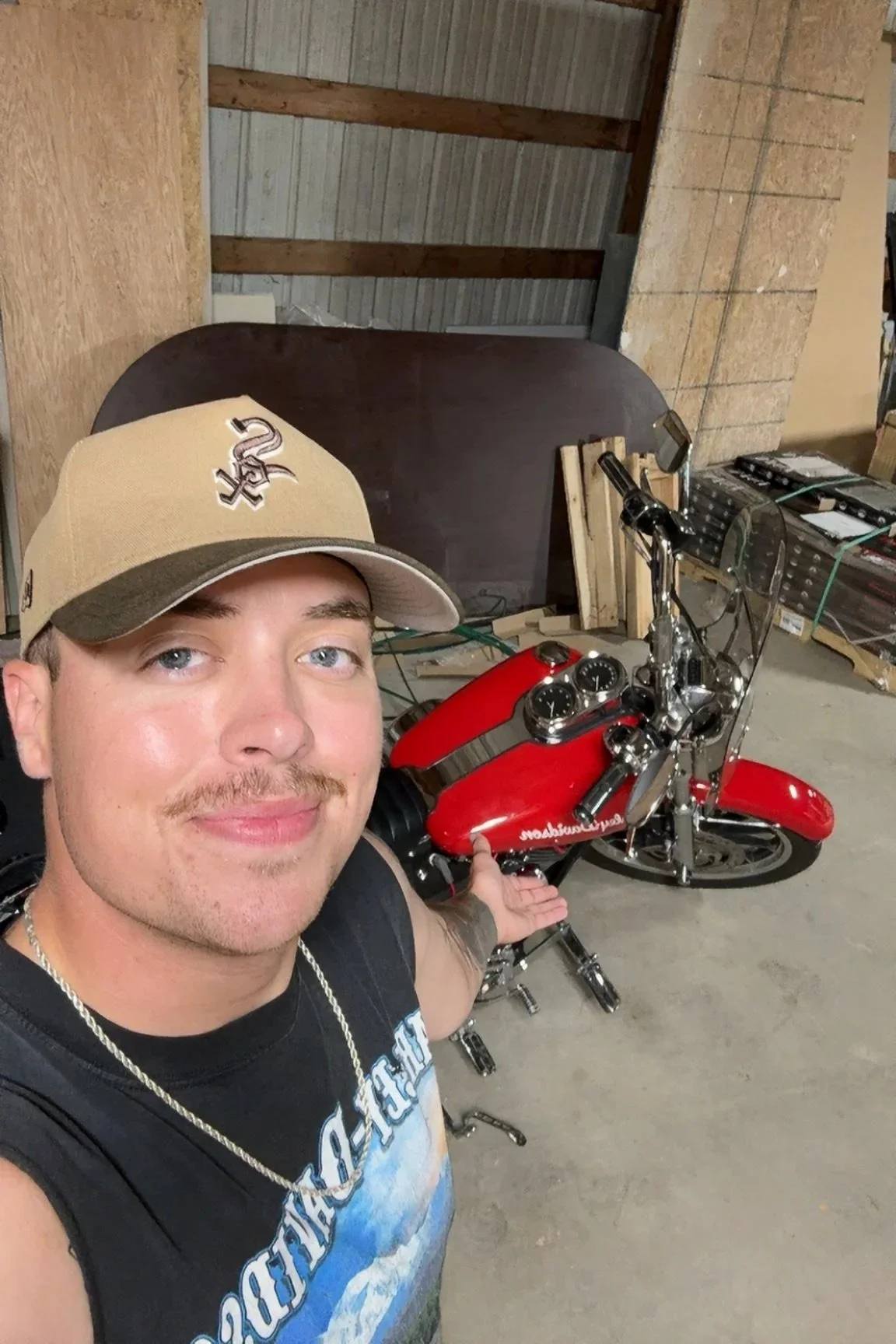 A man taking a selfie inside a garage, pointing at a red motorcycle with chrome accents, a windscreen, and a leather seat, with a smiling expression wearing a beige cap and a black graphic T-shirt.