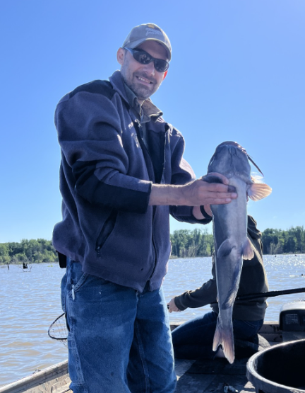 A man wearing sunglasses, a cap, and a fleece jacket holding a large fish he caught during a fishing trip on a boat in a river, with tall grass and trees in the background.