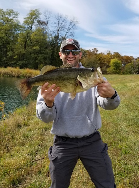 Picture of a man in a gray sweatshirt, hat, and sunglasses holding a fish with a pond and trees in the background.