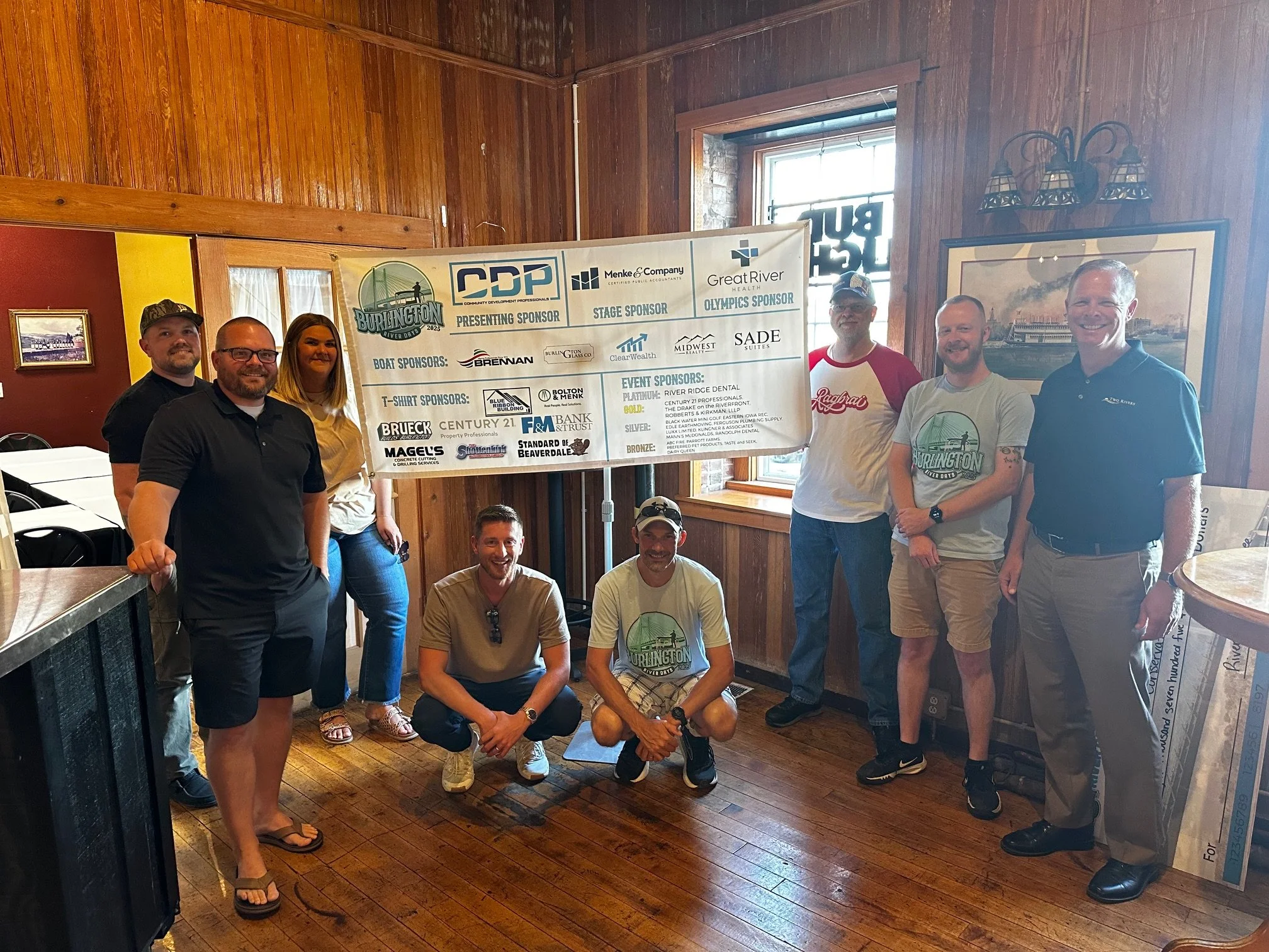 Group of nine people standing and kneeling inside a wooden room, posing for a photo in front of a banner with sponsor logos.