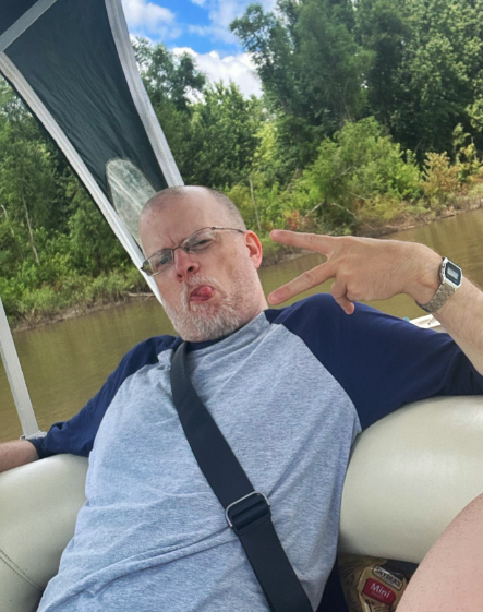 Man with glasses making a peace sign and sticking out his tongue while riding on a boat on a river.
