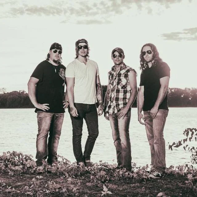 Four men standing outdoors near a body of water with trees in the background, wearing casual clothing and sunglasses, during sunset.