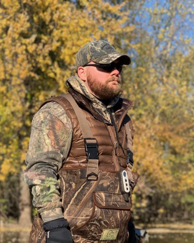 A man wearing camouflage outdoor clothing and sunglasses standing outdoors with fall foliage in the background.