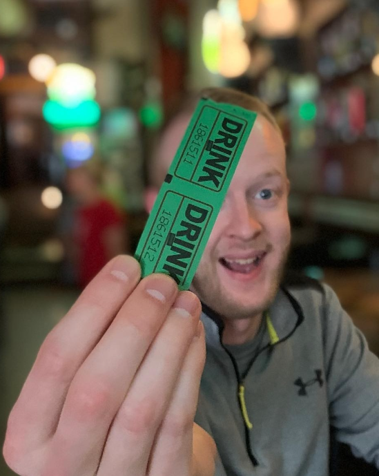 Person smiling and holding two green ticket stubs labeled 'DRINK' in a bar or restaurant setting.